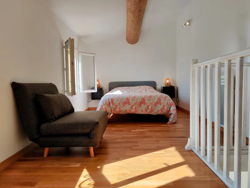 Lit double, fauteuil gris et poutre apparente dans la chambre ensoleillée